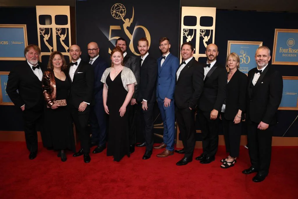 Group of people in formal attire posing on a red carpet at an Emmy awards event, with a backdrop displaying the number 50 and logos.
