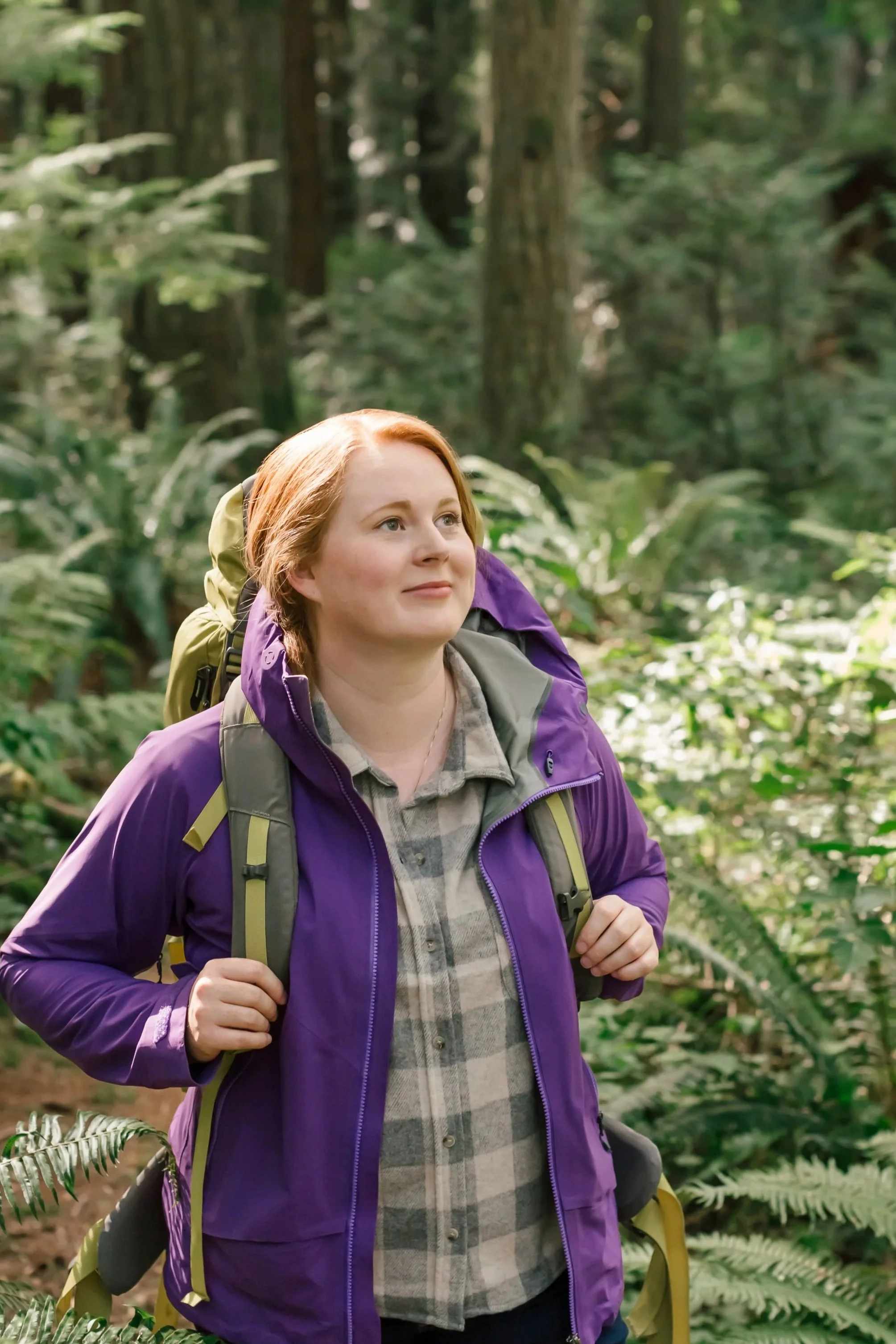 A woman with red hair wearing a purple jacket and backpack hikes through a forest with green foliage.