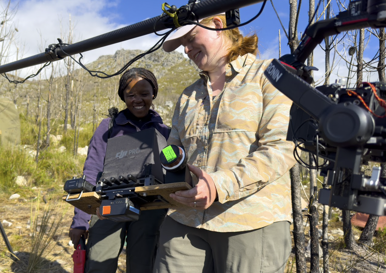Two women outdoors in a natural, mountainous setting with trees and rocky hills, smiling and looking at a drone controller. One woman holds a drone controller with a green indicator screen, and a large camera mounted on a drone is visible in the foreground.