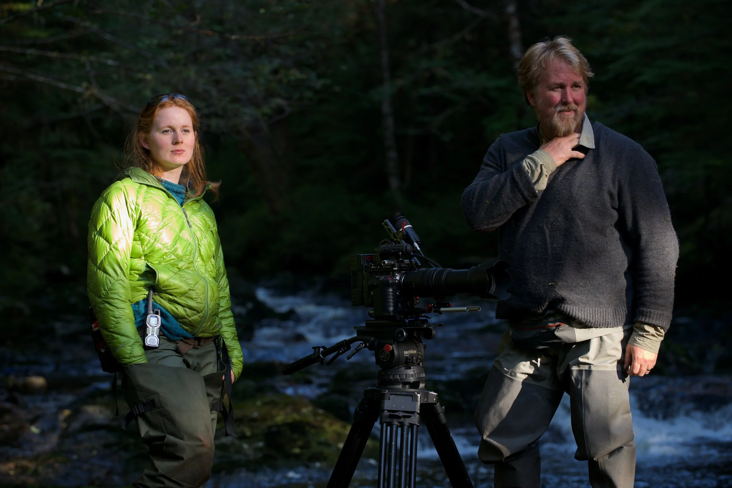 Two people standing outdoors near a flowing stream, with a professional camera on a tripod between them, surrounded by trees.