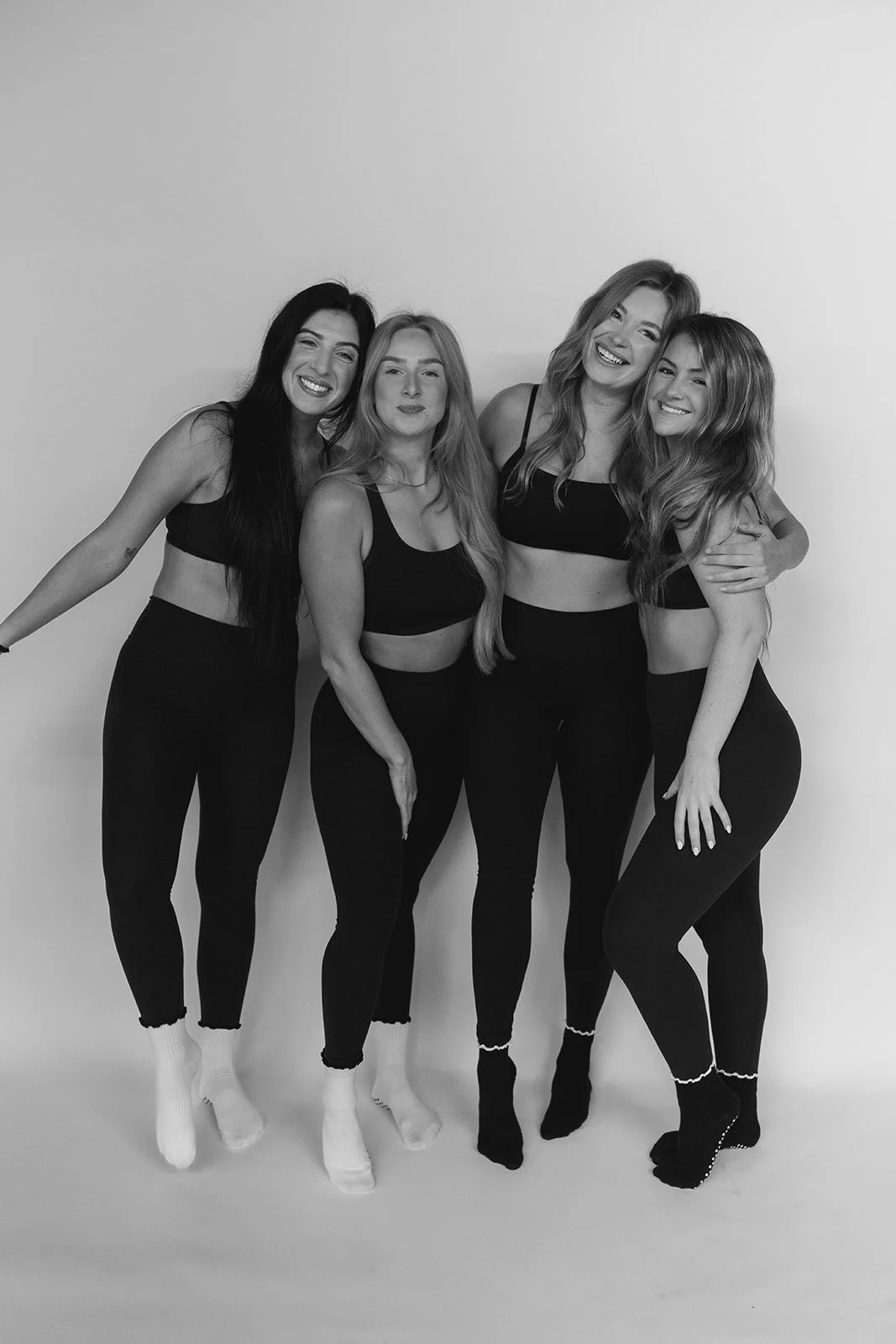 Four women standing together, smiling, dressed in black workout clothes and socks, against a plain wall.