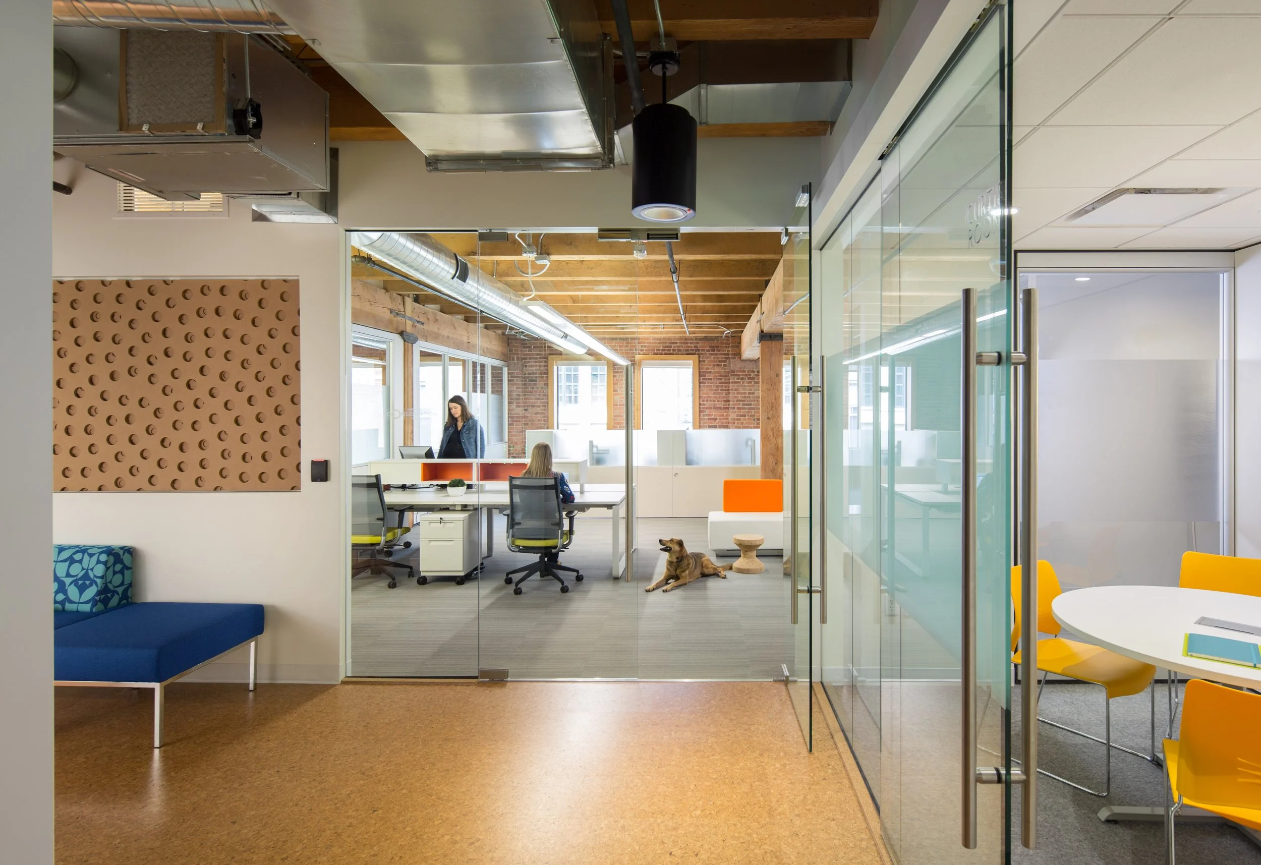 Modern office space with glass walls, yellow chairs, a round white table, and a sitting dog near the glass doors. Office desks and a woman working at her computer are visible in the background.
