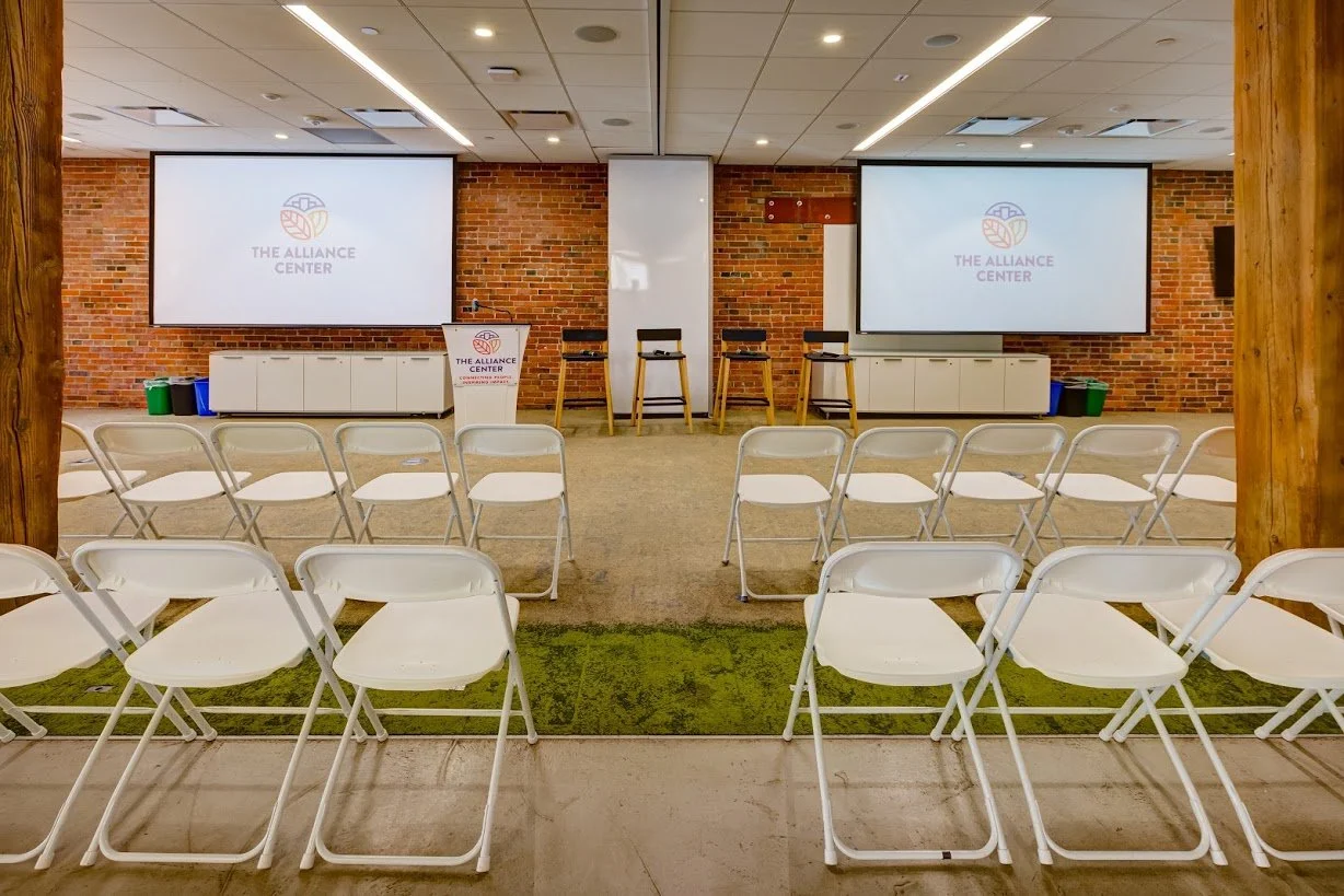 An indoor conference room with a brick wall, two large screens displaying a logo for The Alliance Center, a white podium, three chairs at the front, and multiple rows of white folding chairs facing the screens.