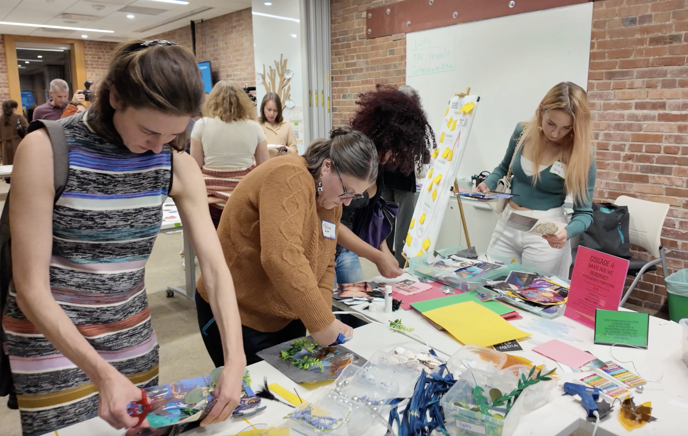 Several women are gathered around a table working on a community art project, with colorful paper, scissors, and decorative items. Others are visible in the background participating in similar activities.