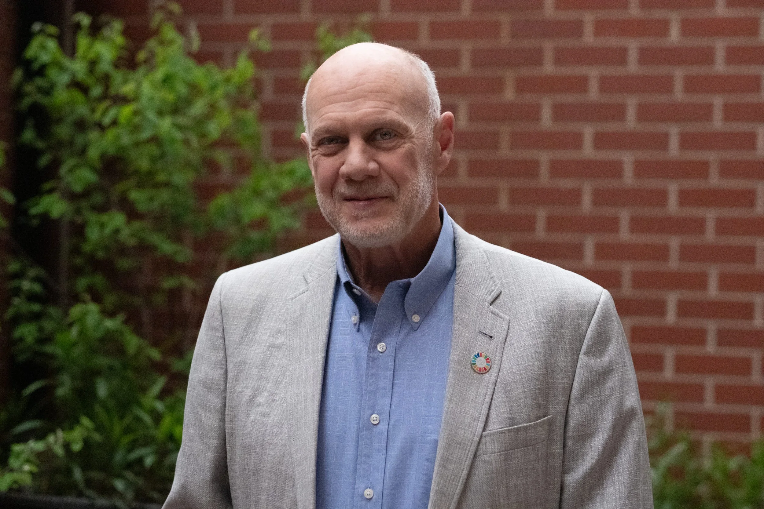 A middle-aged man with a bald head and gray beard wearing a light gray suit jacket over a blue shirt, standing outdoors by a brick wall with green foliage nearby, smiling softly.