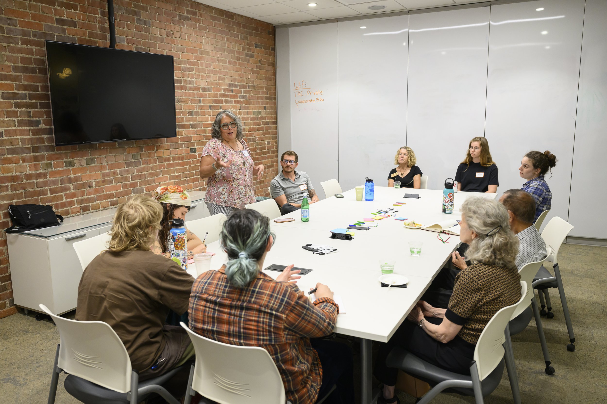 A diverse group of people sitting around a large conference table in a modern meeting room, listening to a woman with curly gray hair and glasses who is speaking. The room has a brick wall on one side, white panels on the other, and a large television screen mounted on the wall.