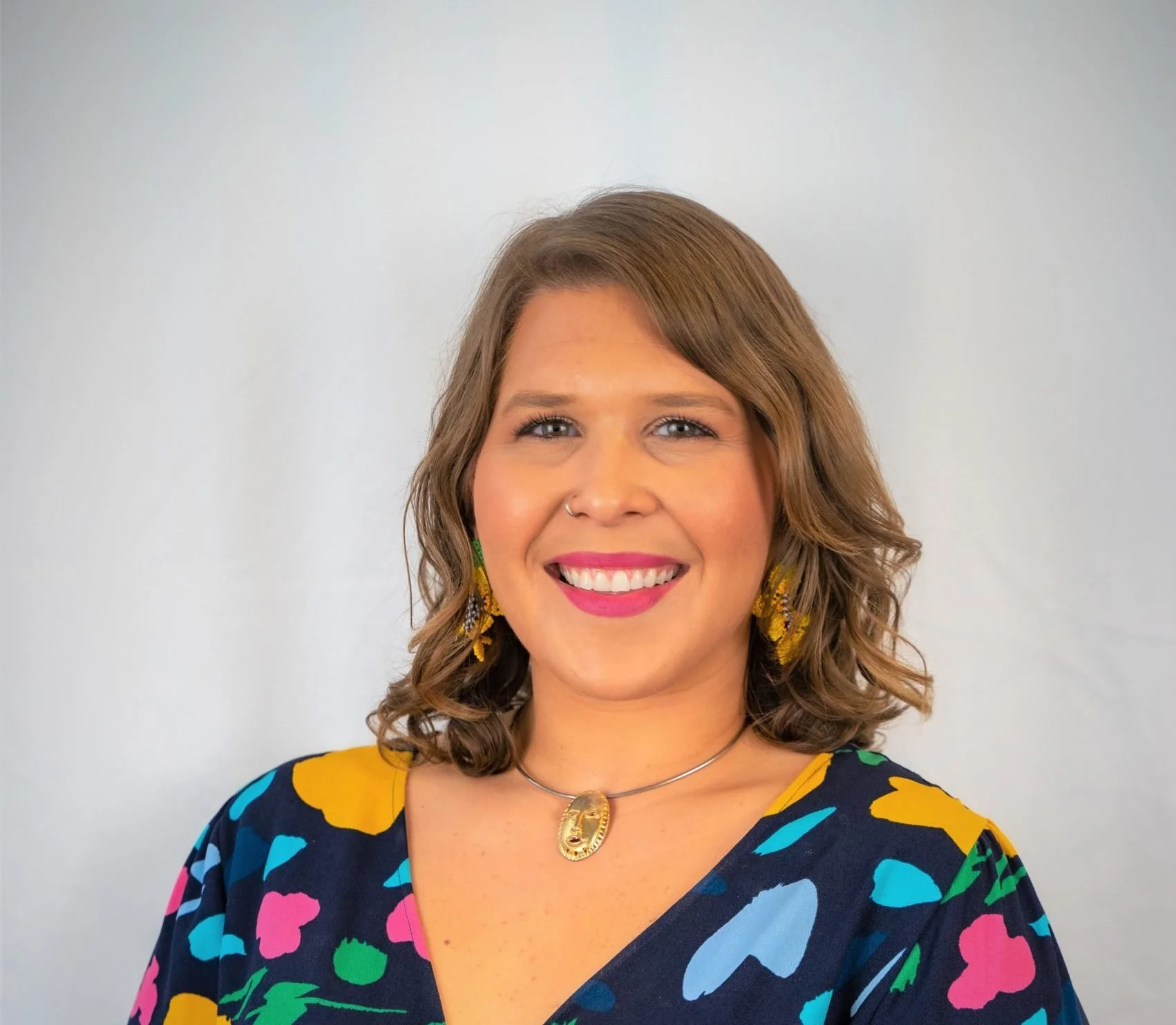 A woman with shoulder-length brown wavy hair, smiling, wearing yellow earrings, a gold nose ring, a colorful floral dress, a gold necklace with a pendant, standing against a plain white background.