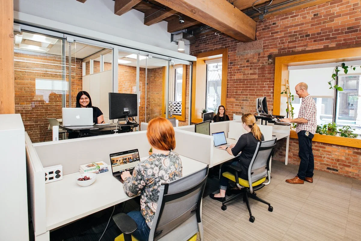 An open-plan office with exposed brick walls, featuring multiple employees working at desks with computers and a man standing and working at a desk. The space has large windows with yellow frames, potted plants, and a wooden ceiling.