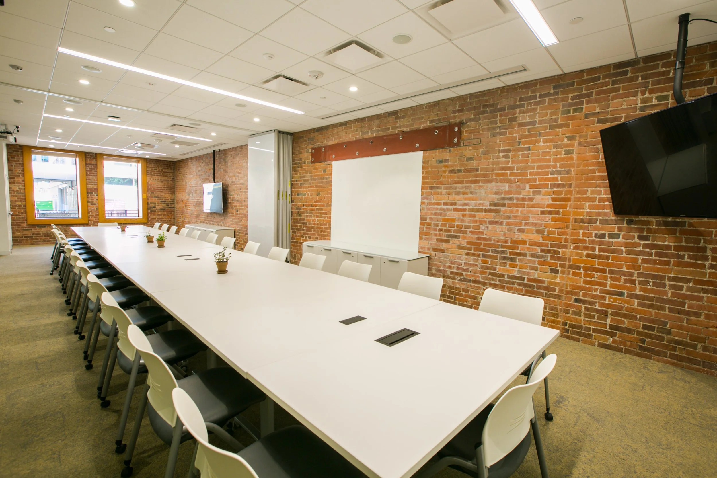 Empty conference room with long white table, white chairs, brick walls, windows, and a mounted flat-screen TV.