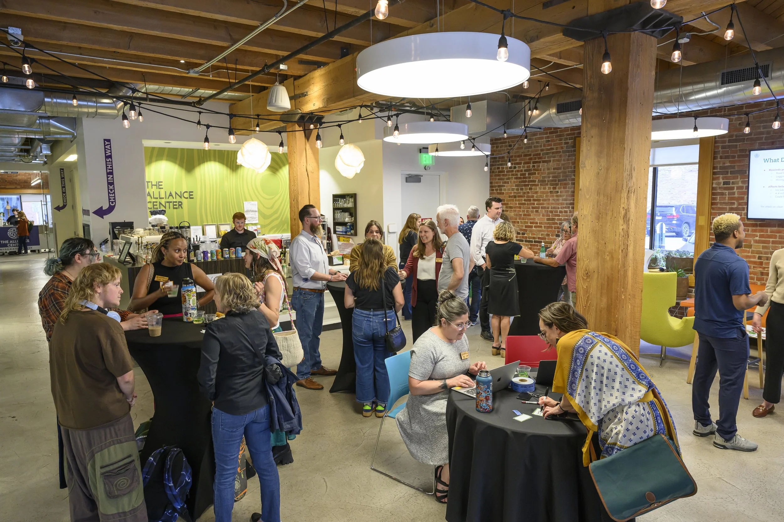A group of diverse people gathered in a modern indoor space with wooden beams, exposed ductwork, and string lights. Some are sitting at tables, while others stand and chat near a bar or registration area.
