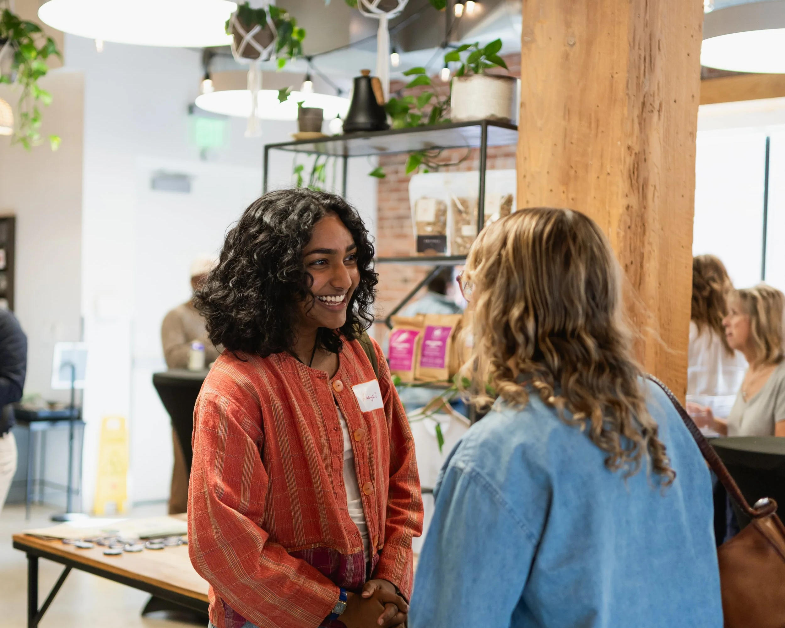 Smiling woman with curly hair wearing a red plaid jacket talking to a woman with straight hair and glasses at a social event in a bright indoor space with wooden beams and plants.