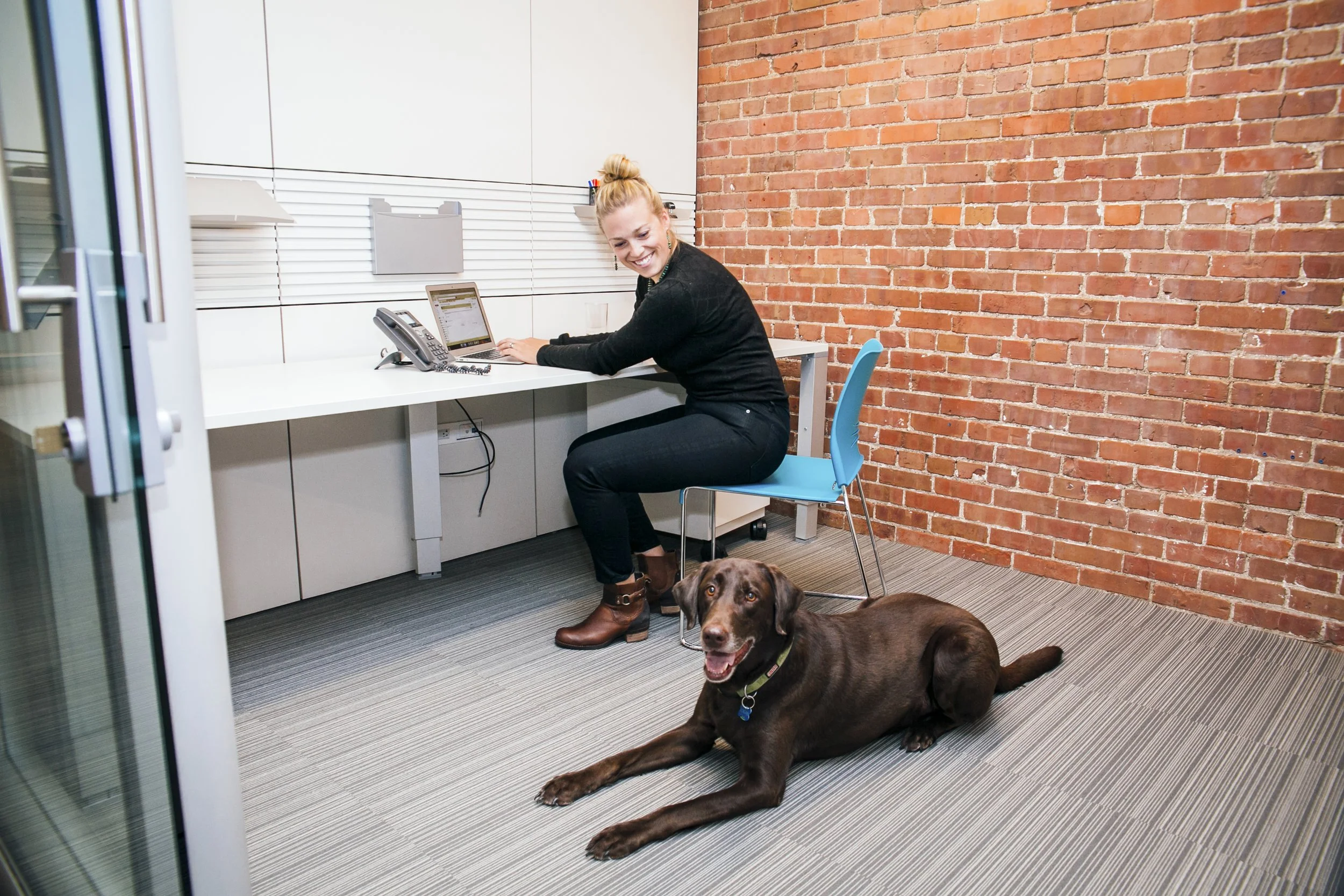 A woman sitting at a white desk in an office with a brick wall, smiling at a laptop. A brown dog lies on the floor nearby, looking at the camera.