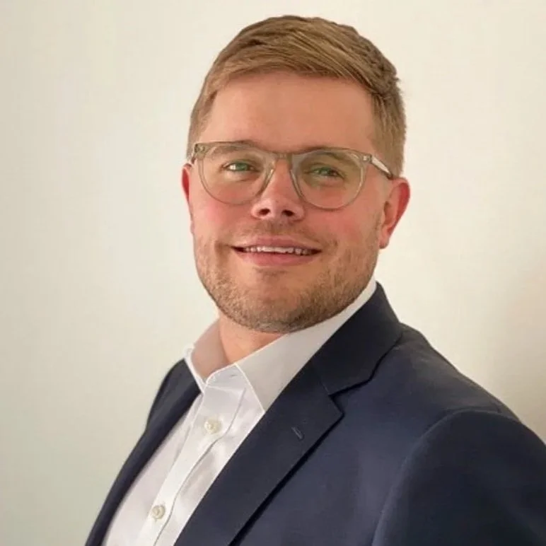 Portrait of a smiling man with short light brown hair, glasses, wearing a dark suit jacket and white collared shirt against a plain light-colored background.