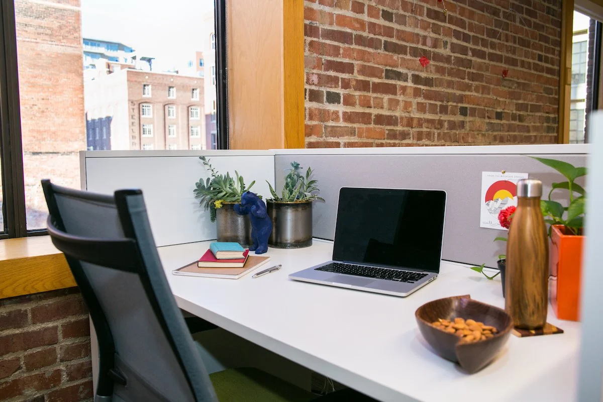 An organized office cubicle with a white desk, black ergonomic chair, two potted succulents, a blue bear figurine, books, a notepad, a pen, a laptop, a copper water bottle, a wooden bowl with snacks, a potted plant, and a colorful wall art in a brick-walled office with large windows showing buildings outside.