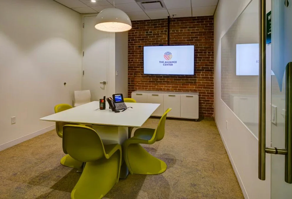 Empty modern conference room with white table, six green chairs, phone, and a large screen displaying 'The Alliance Center' logo. Bright lighting and brick accent wall.
