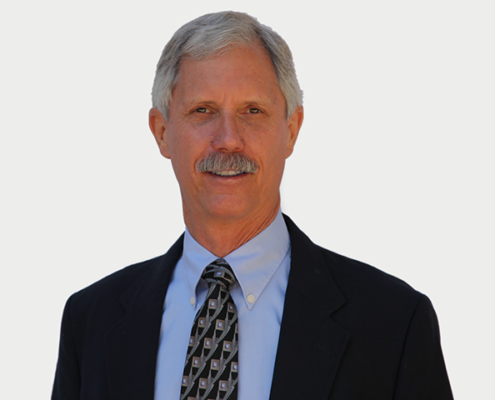 A professional middle-aged man with gray hair and a mustache, wearing a dark suit, light blue dress shirt, and patterned tie, smiling at the camera.