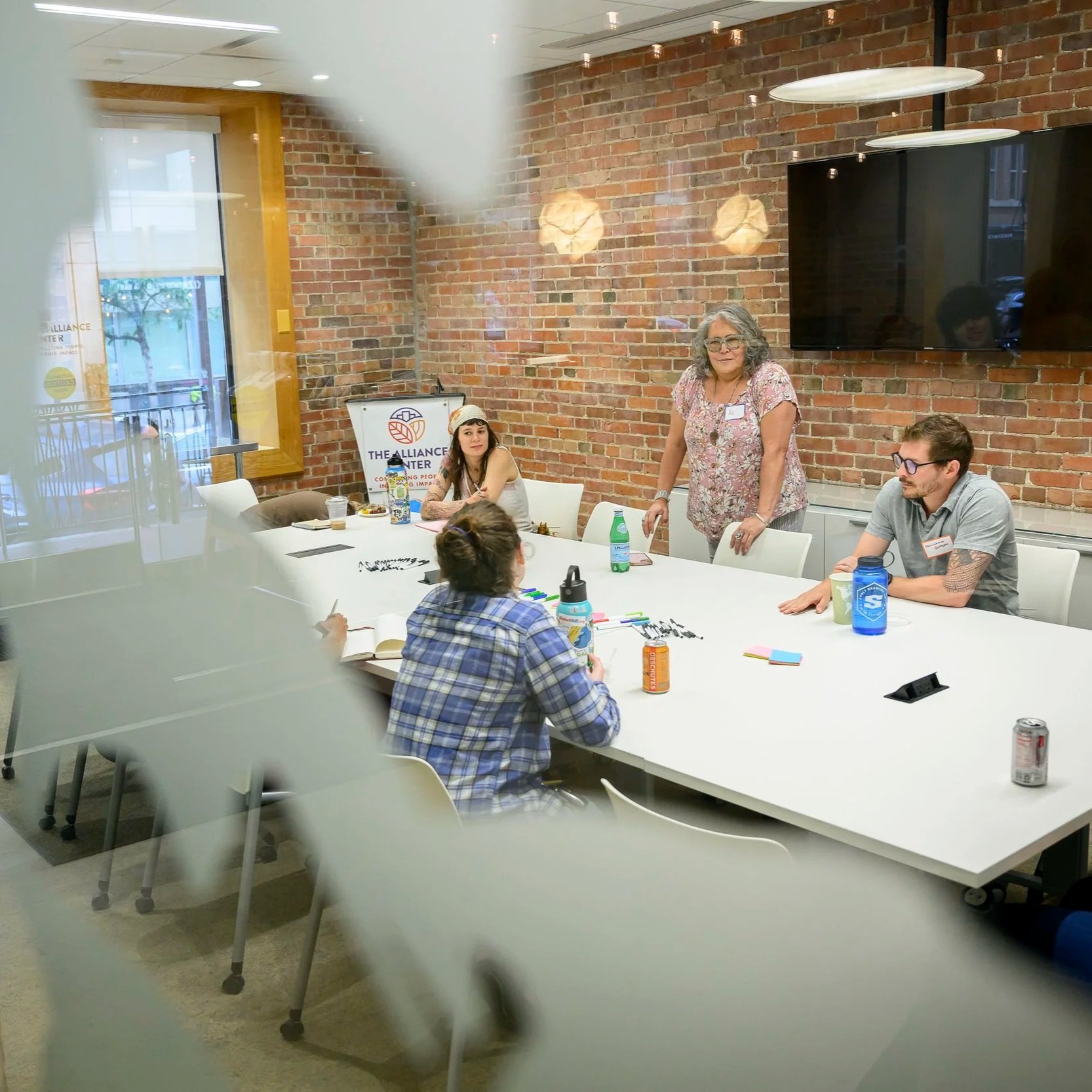 A group of five people sitting and standing around a white conference table in a modern office with brick walls. There is a woman with gray hair standing, speaking or leading a discussion, with name tag. Three others are seated: a woman with long black hair and two men, one with glasses and short hair, and another with tattoos on his arm. The table has notebooks, water bottles, markers, and snacks, with a large flat-screen TV mounted on the brick wall and windows showing an outdoor city scene.