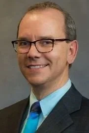A man in glasses, wearing a dark suit, light blue shirt, and blue tie, smiling against a neutral background.