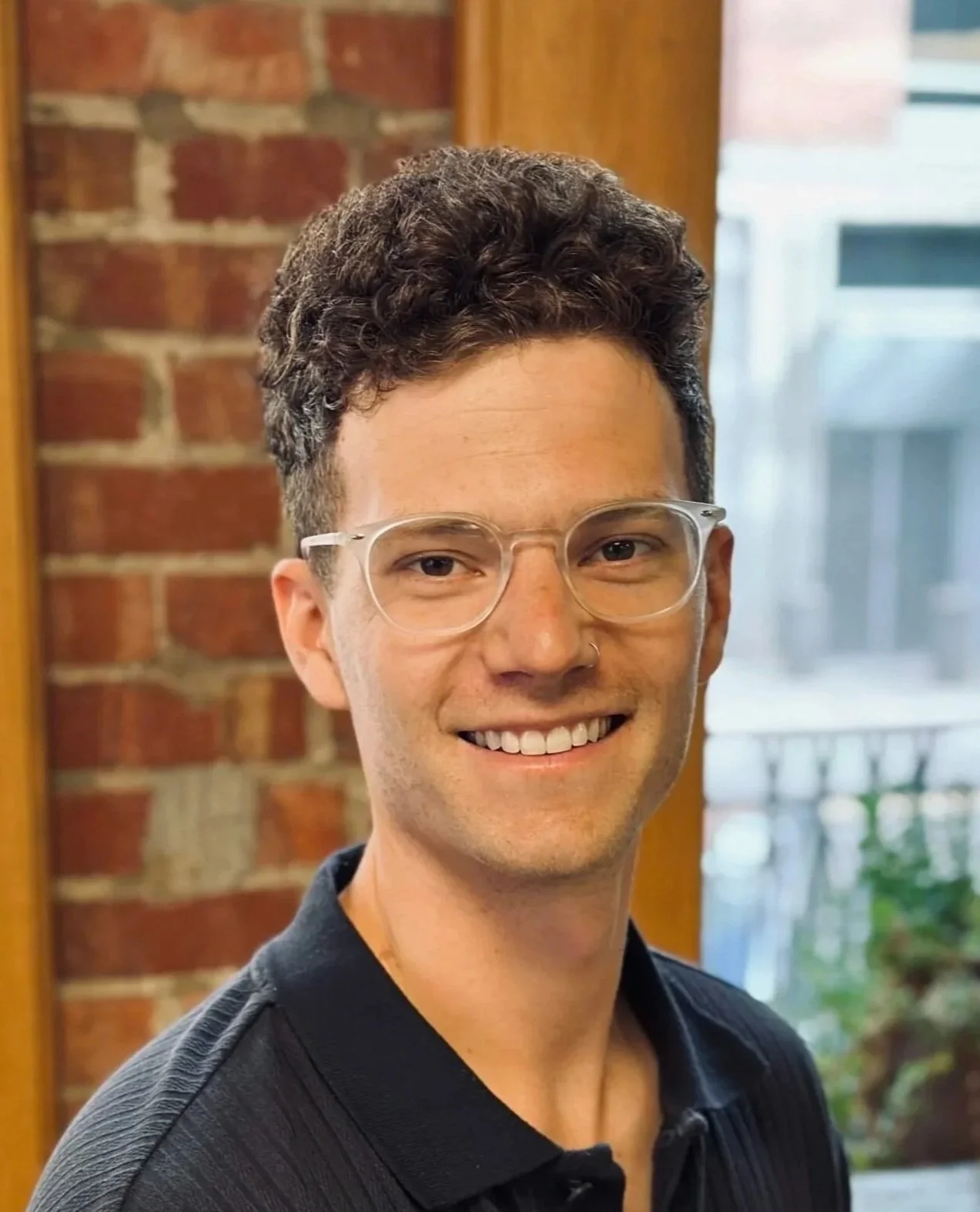 A young man with curly dark hair, glasses, and a nose ring, smiling while standing indoors in front of a brick wall and window.