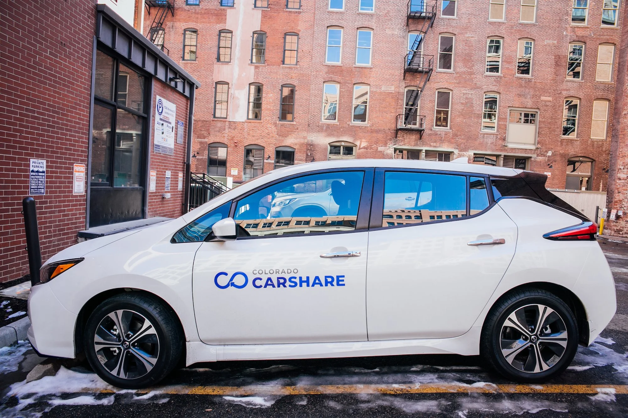 A white Colorado Carshare vehicle parked in an alleyway against a brick building with multiple windows and fire escapes. The vehicle has a logo with a blue infinity symbol and the words "Colorado Carshare." There is snow on the ground and black parking bollards.