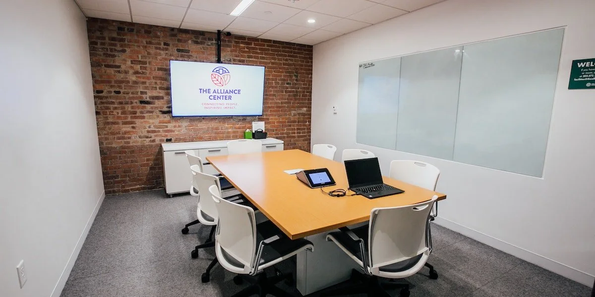 Conference room with a wooden table, six white chairs, a laptop, a tablet, and a whiteboard. A brick accent wall with a TV displaying "The Alliance Center" logo, and a white cabinet underneath.