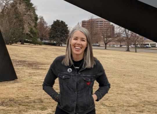 A young woman with gray hair smiling outdoors in a park, wearing a black denim jacket, with trees and buildings in the background.