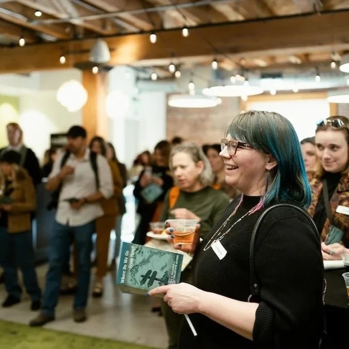 A woman with glasses and blue-green hair smiling while holding a glass of beer and a pamphlet at a crowded indoor event.