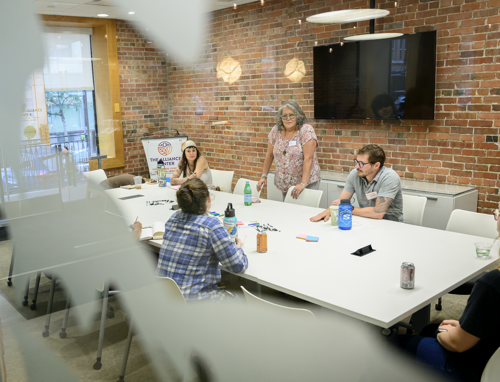 A diverse group of people in a meeting room with exposed brick walls, sitting around a white conference table with notebooks, drinks, and markers, engaged in discussion with a woman standing and speaking.