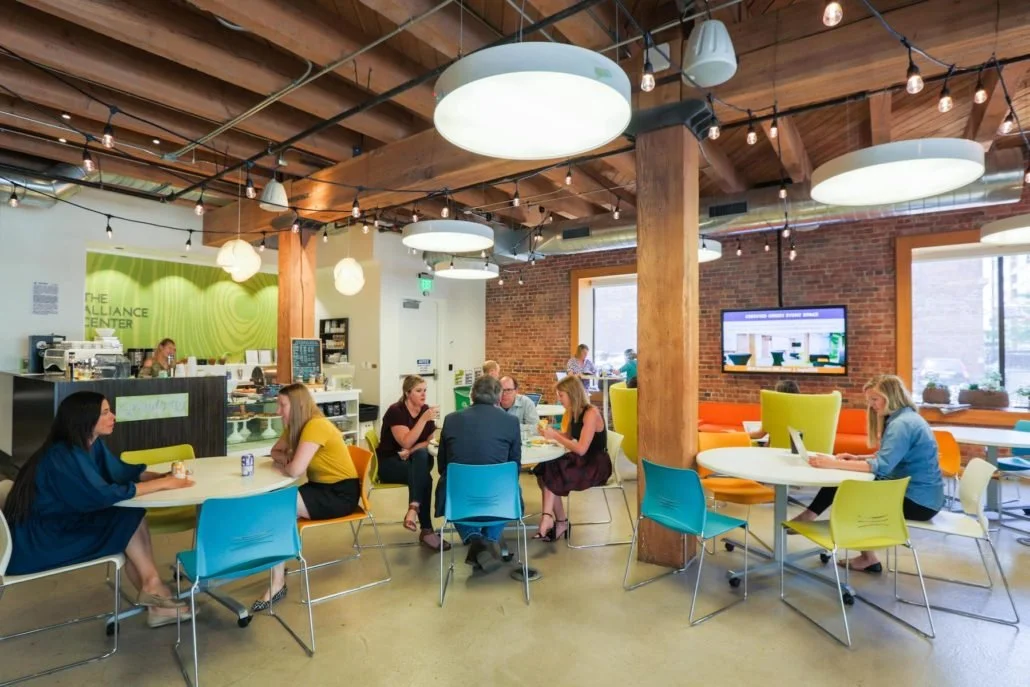 People sitting at tables in a modern, brightly lit cafe with brick walls, wooden beams, and colorful chairs, some using laptops and enjoying drinks.