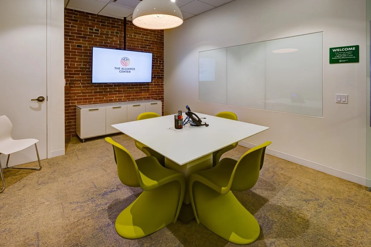 Meeting room with a white table surrounded by six lime green chairs, a brick accent wall with a mounted TV displaying 'The Alliance Center' logo, a white cabinet below the TV, a whiteboard on the wall, and a white door.