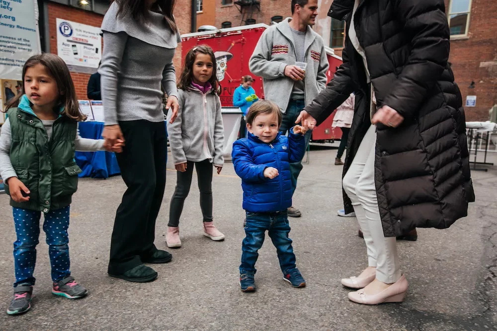 Group of children and adults holding hands outdoors, with one young boy in a blue jacket dancing or playing.