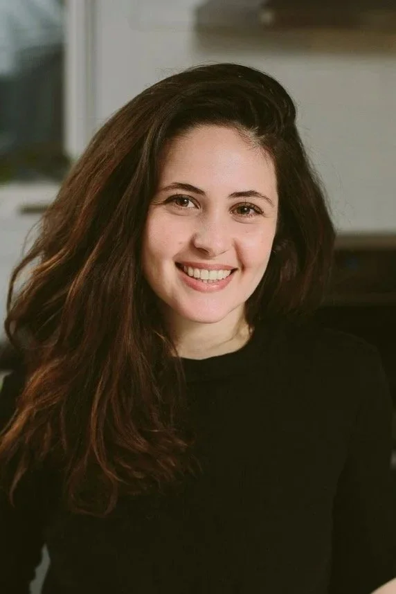 A young woman with long, dark hair smiling at the camera, wearing a black top in an indoor setting.