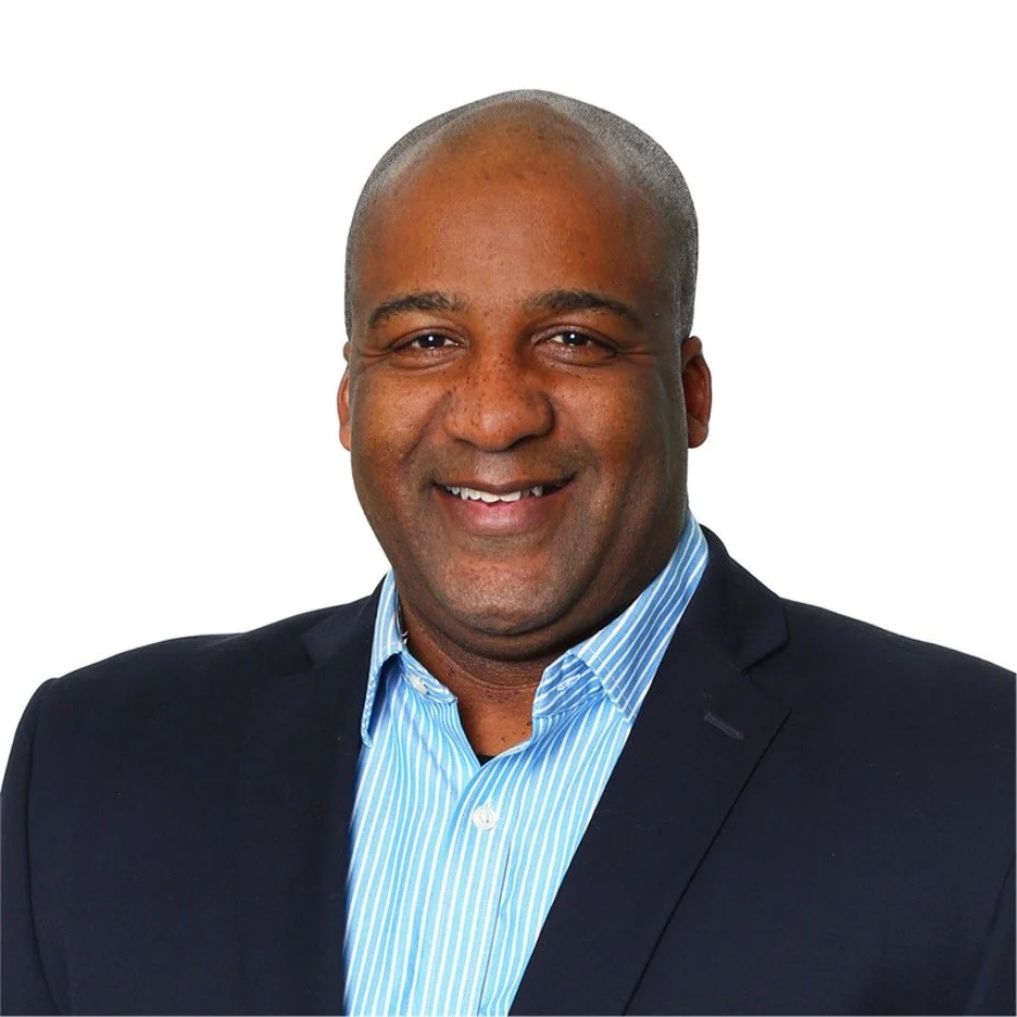 Headshot of a smiling Black man in a dark suit and light blue striped shirt against a white background.