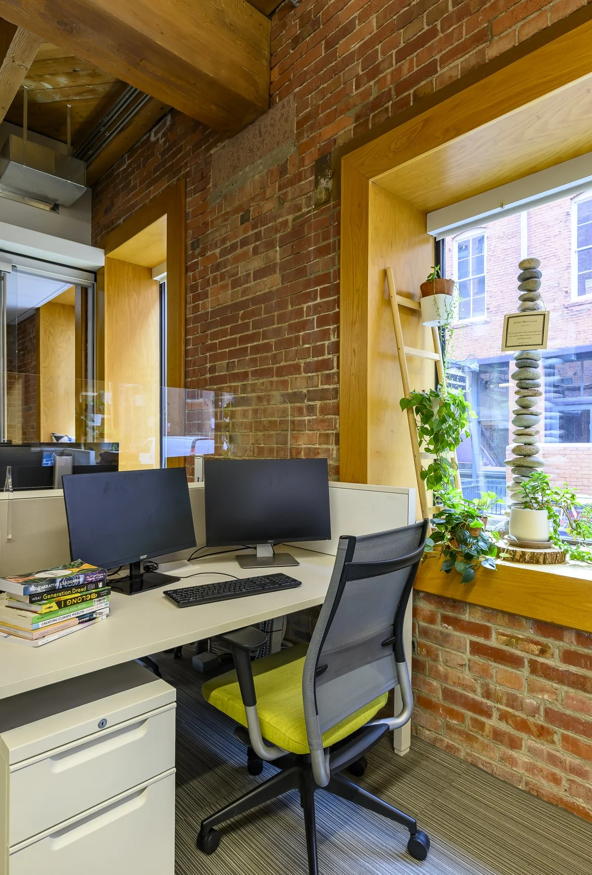 A coworking desk at the Alliance Centerwith exposed brick walls, wooden window frames, two computer monitors, a white desk with books, and a window with potted plants and a stone sculpture.
