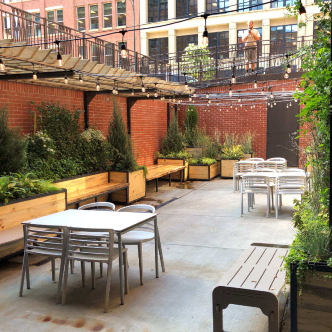 An outdoor patio with several white tables and chairs, wooden planters with green plants along brick walls, string lights hanging overhead, and a man standing on an upper level in the background.