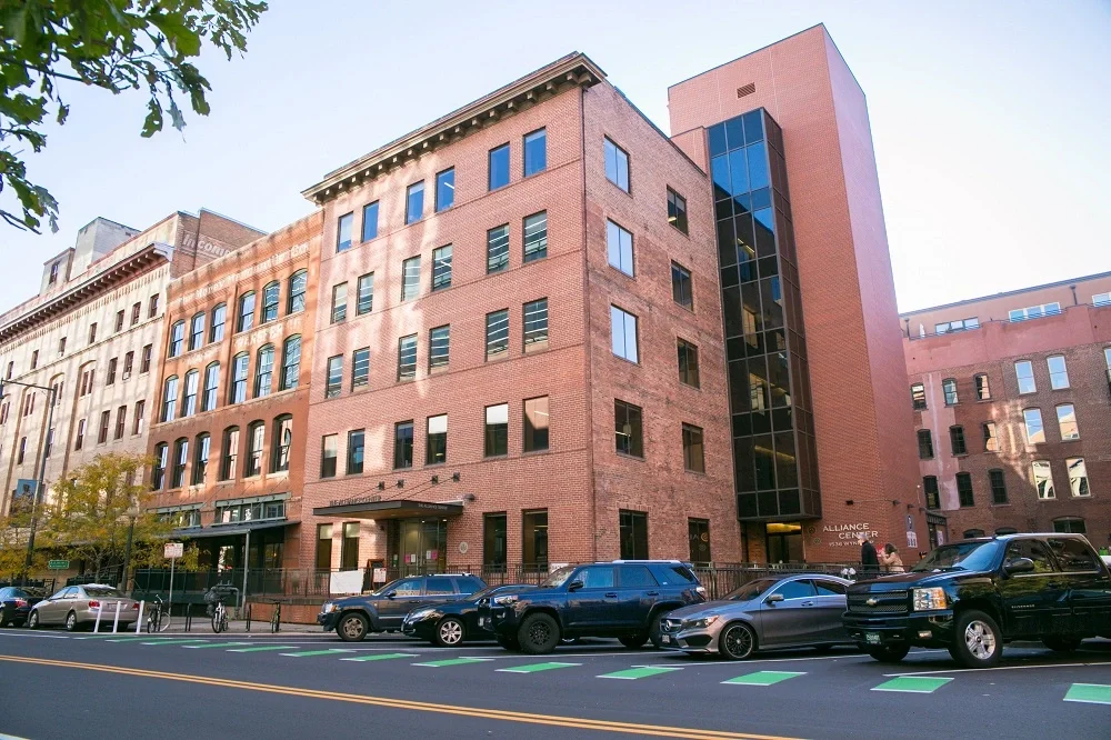 The Alliance Center in lower downtown, Denver. A multi-story brick building with a glass elevator shaft on the right side, next to a street with parked cars and bicycles.