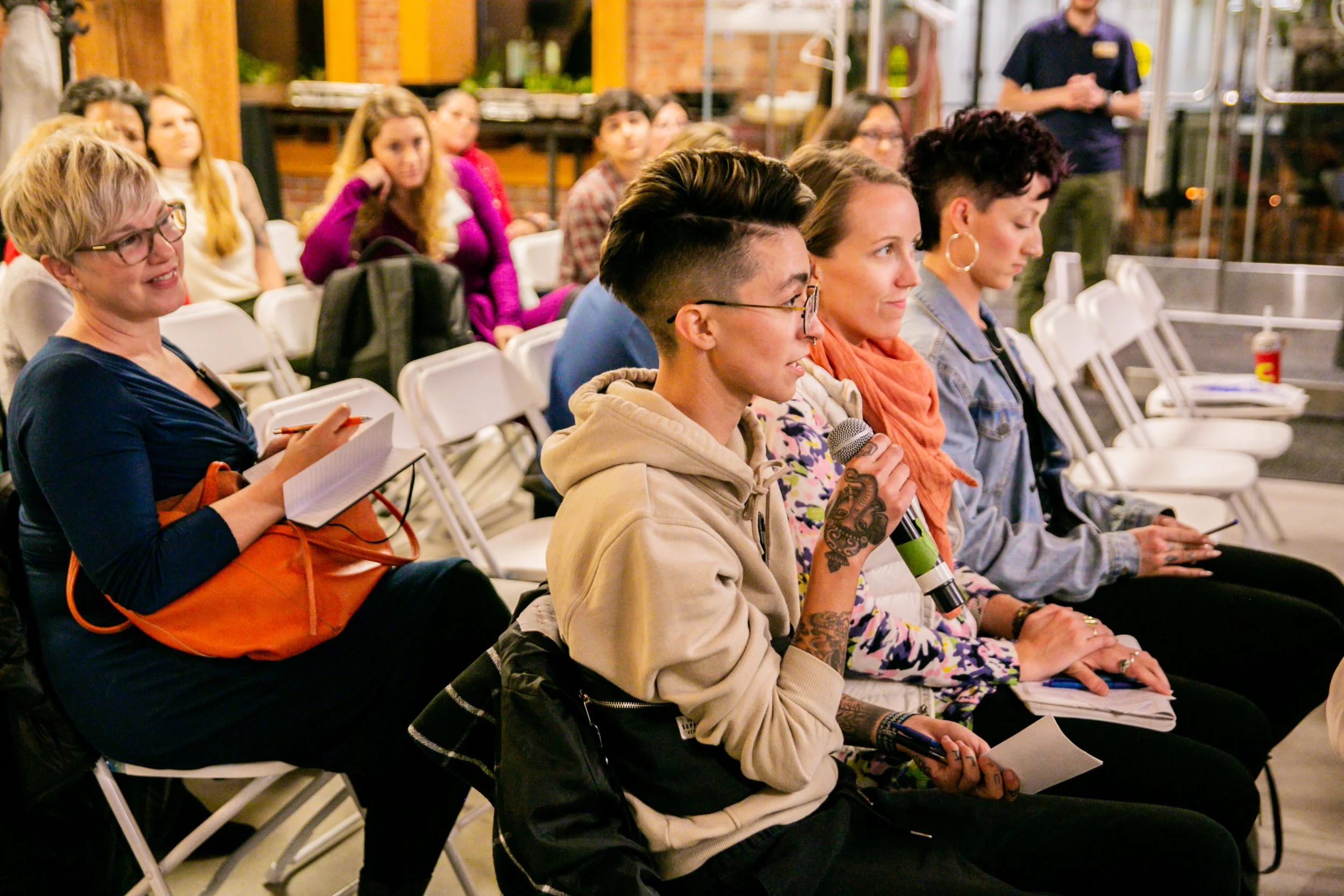 A group of people seated in white chairs attentively listening at an indoor event, with a woman holding a microphone in the center.