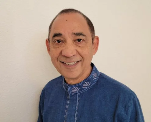 A middle-aged man with short dark hair, smiling, wearing a blue denim shirt with a patterned collar, standing against a plain white wall.
