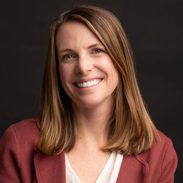 A woman with shoulder-length brown hair smiling, wearing a maroon blazer and a white top, against a dark background.