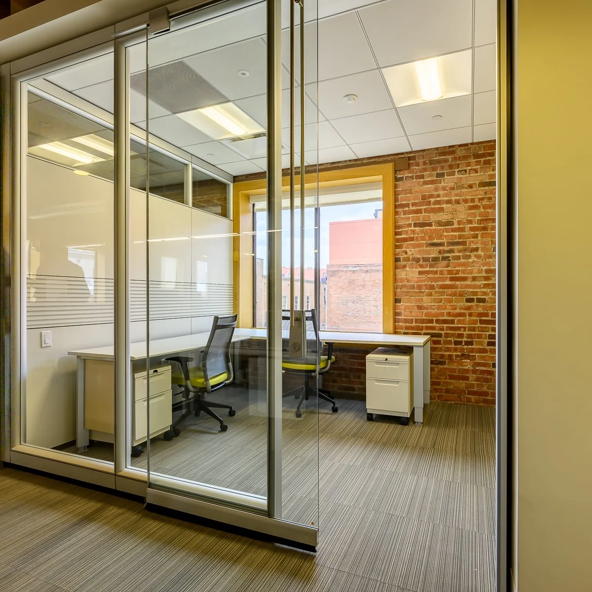 Empty office with two desks, black and green chairs, brick wall, large window, and glass partition.