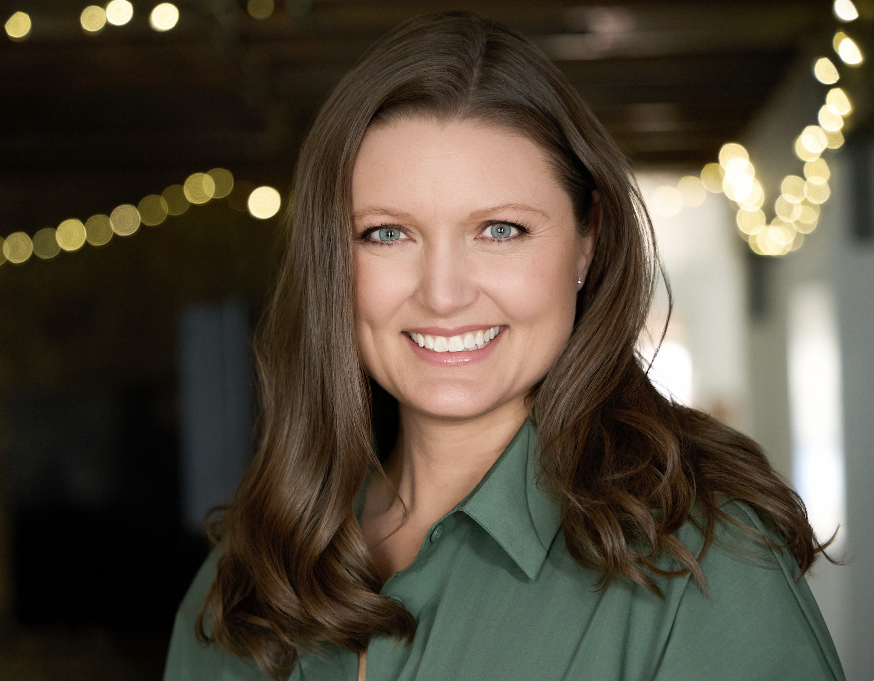 A smiling woman with long brown hair and blue eyes, wearing a green shirt, in a setting with blurred string lights in the background.