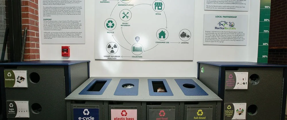 Recycling station with labeled bins for batteries, light bulbs, plastic bags, pet food, foil-lined items, and baby food pouches, located near an informational display about recycling processes.