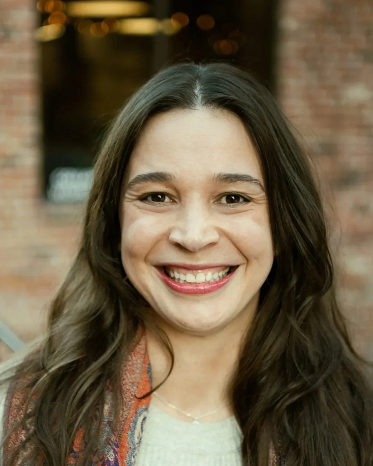 Close-up photo of a smiling woman with long dark hair, wearing a patterned jacket, in front of a brick wall background.