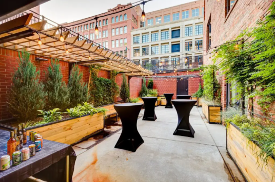 Outdoor urban patio at the Alliance Center with high-top tables, potted plants, and string lights, surrounded by brick.