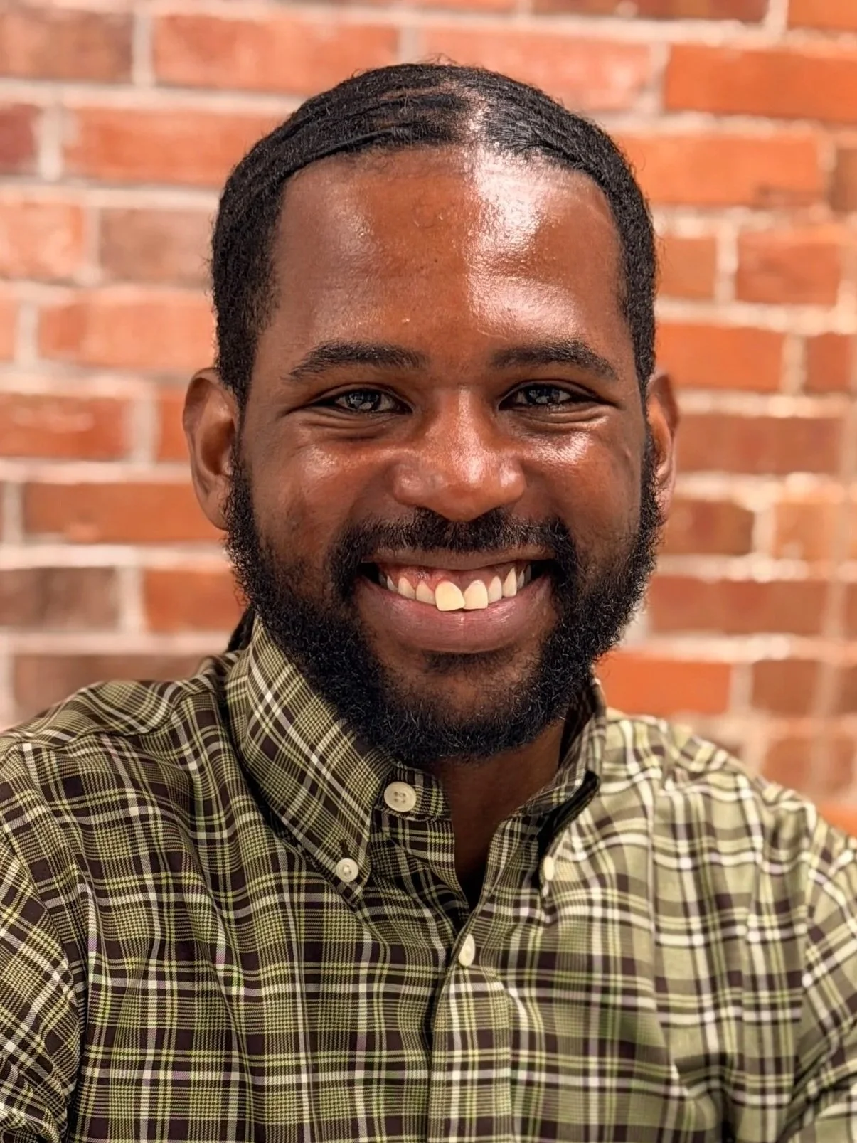 A smiling person with a beard wearing a green plaid shirt in front of a brick wall.