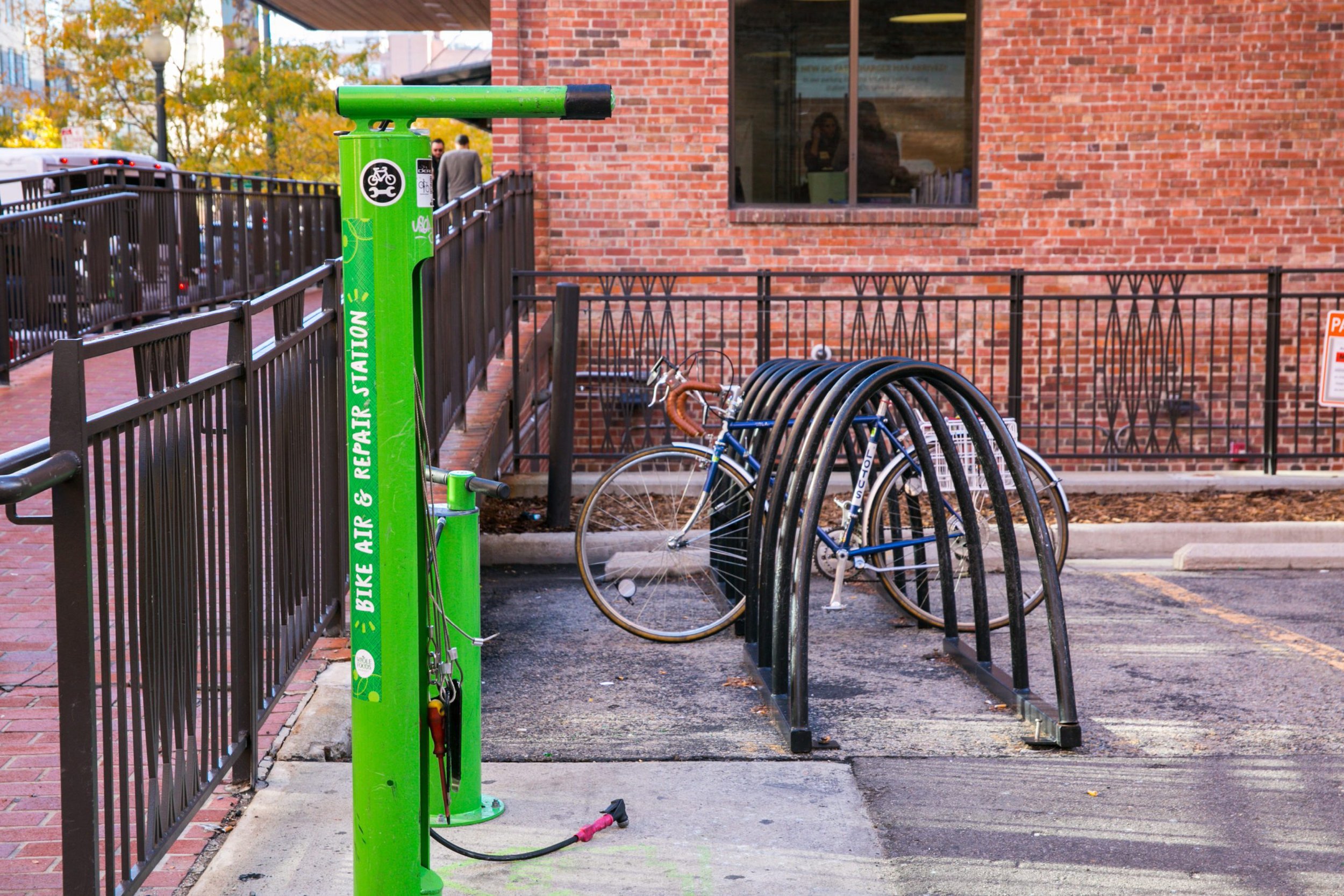 A green bike share station with a bicycle locked to it, a row of bike racks with bicycles, a brick building with a window, and people walking in the background.