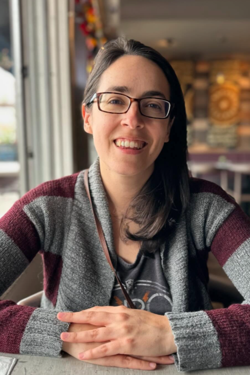 Smiling woman with glasses and dark hair sitting at a table in a cafe, wearing a striped sweater.