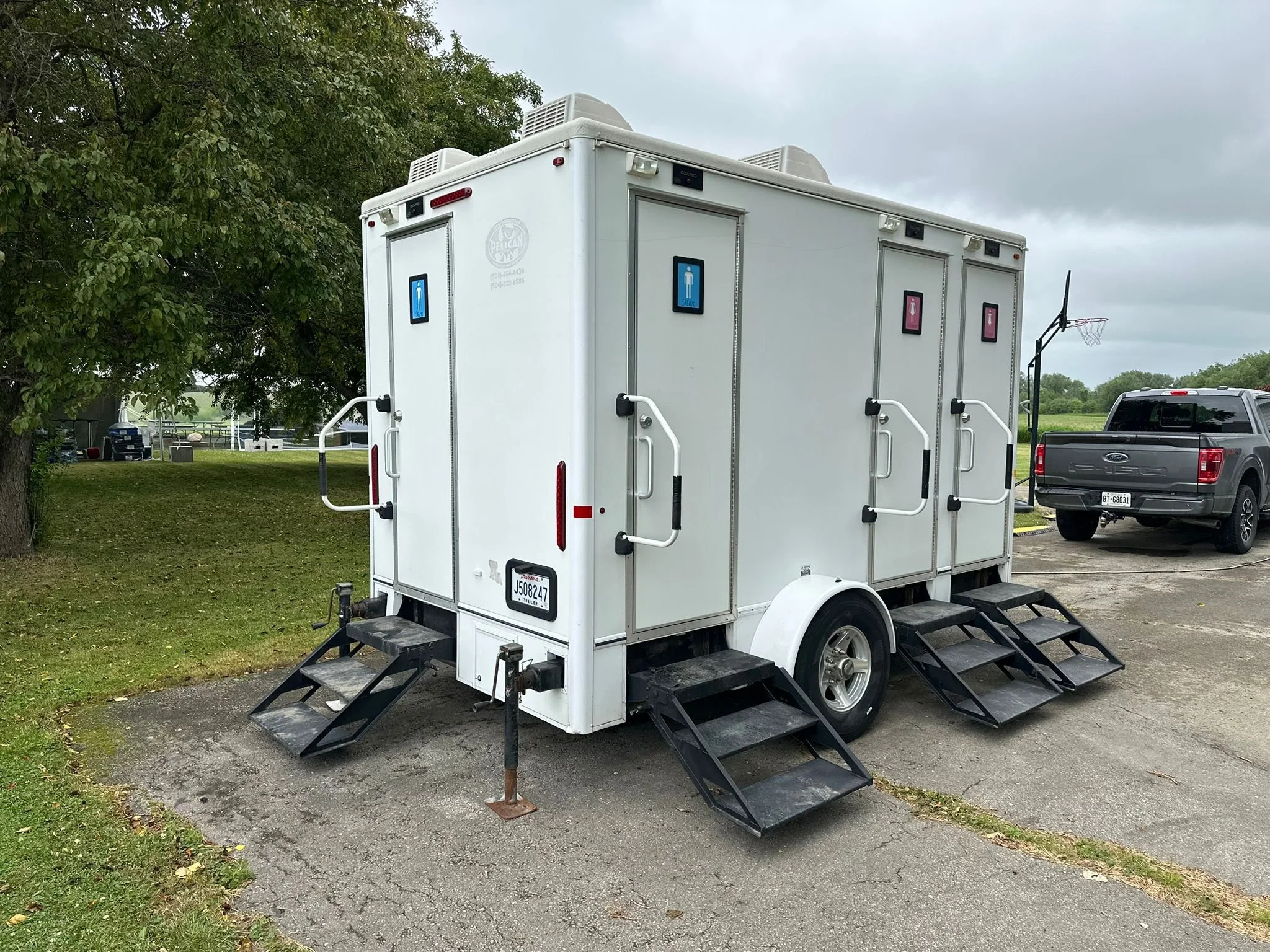 Portable restroom trailer with three separate units, each with steps and signs indicating men's and women's restrooms, parked on an outdoor concrete area near a grassy field and trees.