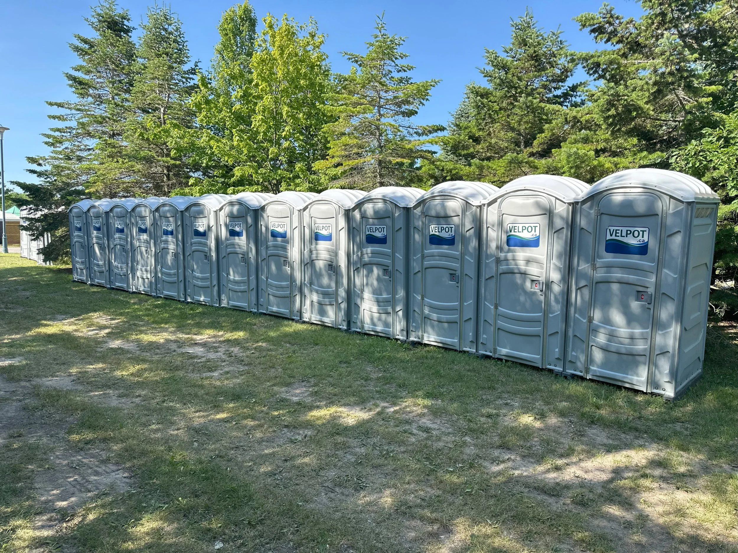 Row of portable toilets with blue and white 'VelpOT' logos on a grassy area with trees in the background.