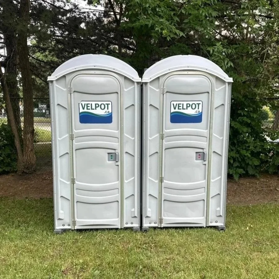 Two portable toilets branded Velpot, positioned side by side on a grassy area with trees and a chain-link fence in the background.